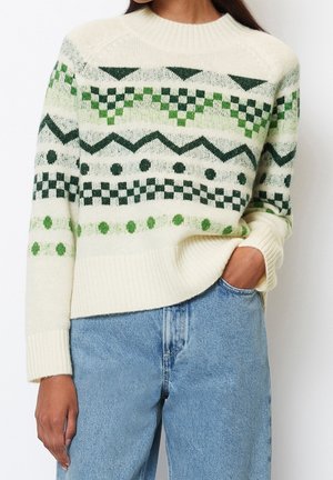 Kvinde iført cremefarvet sweater med grønne geometriske mønstre og lyseblå jeans, hånd i lommen, vist fra skuldre til hofter.