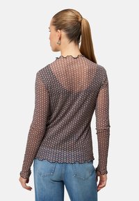 Haut noir transparent à manches longues avec un motif de losanges roses, ourlet festonné et poignets à volants, porté avec un jean bleu.
