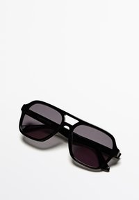Schwarze rechteckige Sonnenbrille mit dunkel getönten Gläsern, auf einer weißen Oberfläche liegend.
