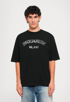 Giovane con capelli ricci che indossa una t-shirt nera oversize DSQUARED2 Milano e jeans blu scoloriti, in piedi davanti a uno sfondo bianco semplice.