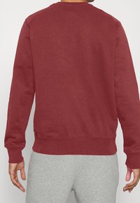 Mörkröd sweatshirt med ribbad krage, långa ärmar och ribbade muddar och fåll. Slät textur, sedd bakifrån, i kombination med grå byxor.