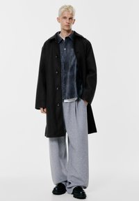 Zwarte wollen jas, donkerblauw overhemd met knopen, grijze joggingbroek, zwarte zware schoenen. Model staat op een effen witte achtergrond.