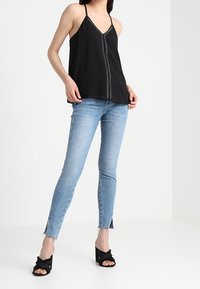 Top nero senza maniche con scollatura a V e cuciture decorative, abbinato a jeans skinny blu chiaro con orli sfrangiati e sandali neri con tacco alto.