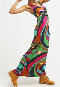 Sukienka maxi w intensywnych wzorach w odcieniach zieleni, różu, pomarańczy i czerni; bezrękawowy fason z dopasowaną talią i paskiem w akcentującym kolorze. Noszona z żółtymi botkami.