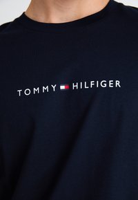 Marineblå bomulls T-skjorte med hvit "TOMMY HILFIGER" logo med en rød og hvit flaggdetalj i midten. Korte ermer og rund hals design.