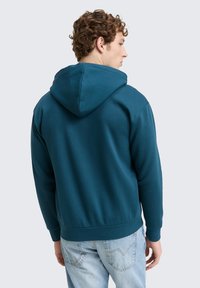 Sudadera con capucha de color azul verdoso con una textura suave, puños y dobladillo acanalados. Marcas de la casa en un bolsillo trasero de unos vaqueros de color azul claro.