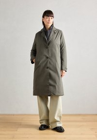 Olijfgroene, geknielengte jas met een gestructureerd ontwerp, knoopsluiting en zijzakken. Draag het over een grijs kledingstuk en gecombineerd met een broek met wijde pijpen.