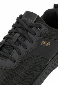 Svarta sportskor med en strukturerad mesh-överdel, släta gummidetaljer, snörning och en guldpräglad "BOSS"-logotyp på sidan.