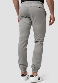 Joggers grises con puños elásticos, que presentan bolsillos laterales, un bolsillo con cremallera en la parte posterior y una tela suave y texturizada. Llevados con zapatillas blancas.