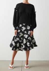 Czarna bawełniana bluza z perłowymi i kokardkowymi akcentami, zestawiona z czarną spódnicą w kształcie klepsydry, zdobioną białymi kwiatowymi wzorami, oraz białymi sandałami.