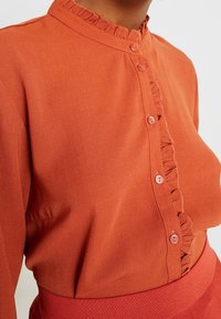 Rustfarbene Bluse mit hohem Kragen, Rüschenbesatz und Knopfverschluss, der mit passenden Knöpfen versehen ist. Der Stoff hat eine glatte Textur.
