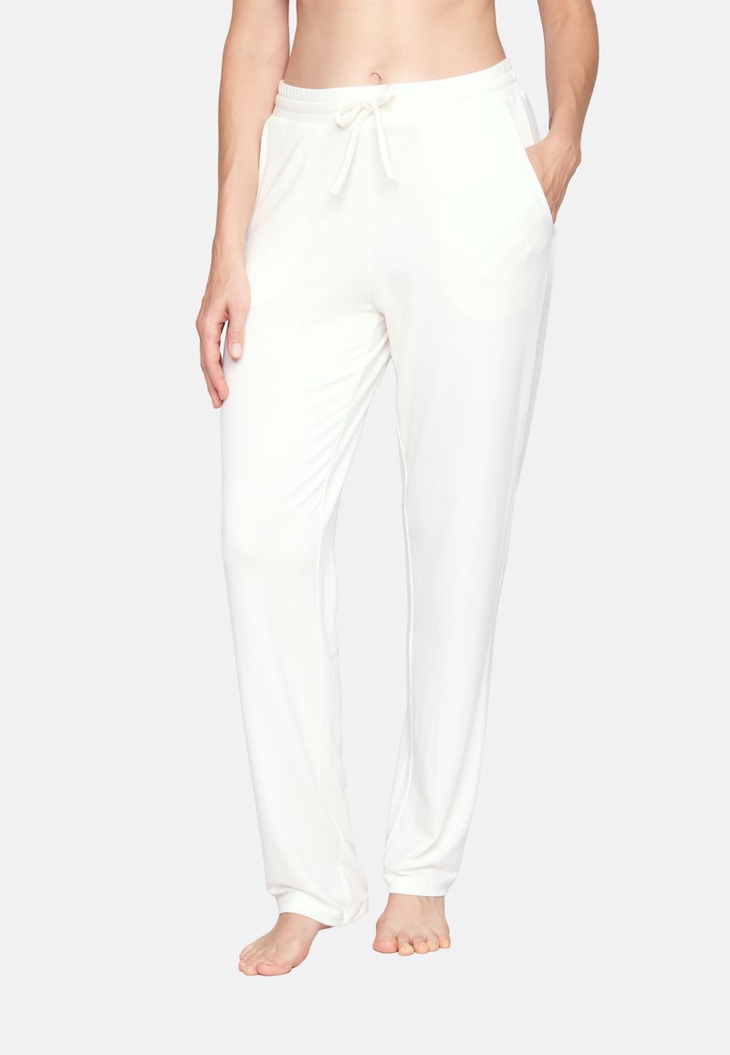 Kobieta w białych spodniach dresowych ze ściągaczem i bocznymi kieszeniami, stojąca boso na jednolitym tle.