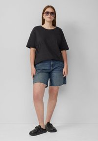 Zwarte katoenen t-shirt met een relaxte pasvorm, gecombineerd met blauwe denim shorts van kniehoogte met een rafelige zoom. Zwarte instappers en een zonnebril.