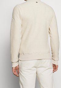 Sweatshirt in pile beige, con texture, con colletto e polsini a costine; presenta un logo discreto sul retro. Abbinato a pantaloni chiari.