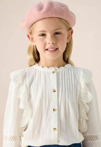 White long-sleeve blouse with ruffled lace trim, gold buttons, and textured fabric. Paired with a soft pink beret.