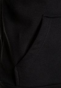 Primo piano di un tessuto di felpa nera, caratterizzato da una texture morbida, una tasca sulla parte frontale e dettagli di cucitura visibili. Il materiale appare resistente.