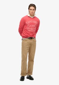 Felpa rossa con grafica bianca, chinos beige e scarpe nere. Outfit casual con colletto a strati e orli dei pantaloni arrotolati.