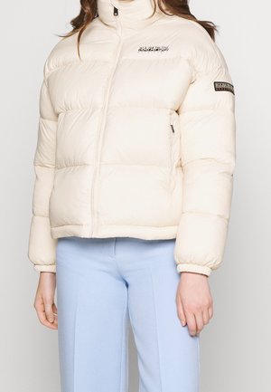 Femme portant une doudoune couleur crème avec fermeture éclair à l'avant et écussons avec logo sur la poitrine et la manche, associée à un pantalon bleu clair.