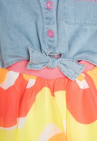 Denimová blůza s růžovými třpytivými knoflíky, uvázaná v pase. Sukně má splývavou tkaninu v oranžové a žluté barvě s růžovým elastickým pasem.