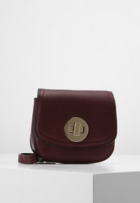 Sac en cuir bourgogne à bandoulière avec un rabat arrondi et un détail en métal circulaire doré. Texture lisse et accents de couture subtils.