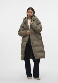 Manteau long matelassé en vert olive, doté d'une capuche, d'une fermeture éclair sur le devant et de poignets élastiqués. Porté sur un jean et des chaussures à bout pointu.