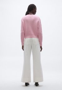 Rózsaszín bordás kötött pulóver kosármintás ujjakkal, fehér bőszárú nadrággal párosítva. A pulóver hátulján egy apró arany díszítés található.