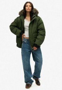 Green puffer jacket with a fur-lined hood, paired with a light gray top and wide-leg blue jeans. Brown lace-up footwear completes the outfit.