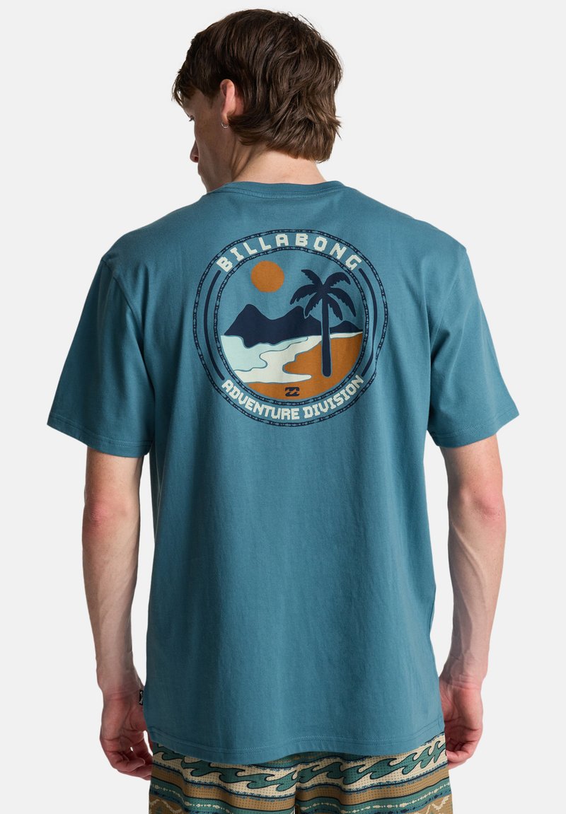 Muž v tyrkysovom tričku Billabong s kruhovým plážovým logom zobrazujúcim palmou, slnko a hory, v kombinácii s nohavicami s potlačou.