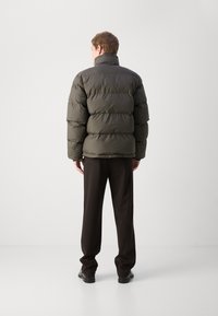 Jachetă pufosă de culoare verde închis, cu un model matlasat orizontal, guler înalt și manșete elastice, asortată cu pantaloni și pantofi negri.