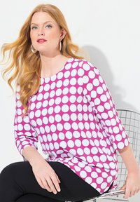 Maglietta lunga viola a maniche lunghe con grandi pois bianchi, scollatura rotonda e vestibilità ampia. Indossata con pantaloni neri, seduta su una sedia di filo.