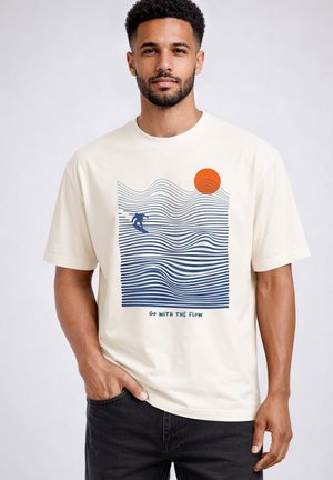 Mann trägt cremefarbenes T-Shirt mit blauen wellenförmigen Linien, Silhouette eines Surfers, orangefarbene Sonne und Text "Go with the flow" auf der Vorderseite.
