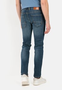 Jean en denim slim fit dans une teinte bleu foncé avec une texture lisse, présentant des poches arrière et une étiquette de ceinture en cuir marron. Assorti avec des chaussures blanches.