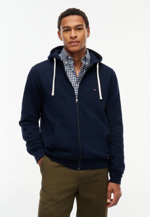 Homme portant un sweat à capuche bleu marine zippé sur une chemise à carreaux bleue et blanche avec un pantalon vert olive, debout avec une main dans la poche.