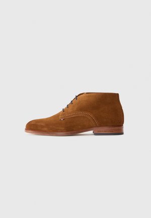 Botas chukka de ante marrón con punta redondeada, cordones azules, sutil detalle de costura y un logo en la suela de cuero.