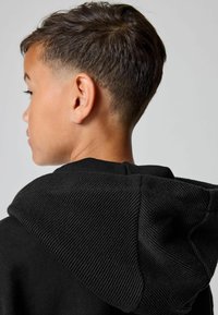 Chico con cabello corto y oscuro, vistiendo una sudadera con capucha negra, mostrado de espaldas y de lado contra un fondo claro y sencillo.