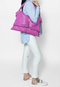 Borsa in pelle rosa con un design annodato, di grandi dimensioni e dalla superficie liscia, indossata sulla spalla. Modello con camicia azzurro chiaro e pantaloni bianchi.