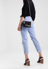 Žena v světle modrých svetších džínách, černých podpatkových sandálech, černém svetru a malé černé křížové tašce s květinovou výšivkou.