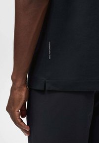 Camiseta de rendimiento negra con abertura lateral, que presenta una tela texturizada y un pequeño detalle de marca en el dobladillo, mostrada en una muñeca y mano.