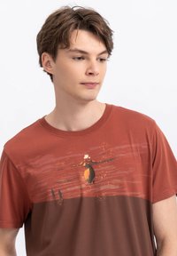 Roestkleurig katoenen T-shirt met een grafische print van een figuur en abstracte achtergrond. Korte mouwen, ronde halslijn en een gladde textuur.