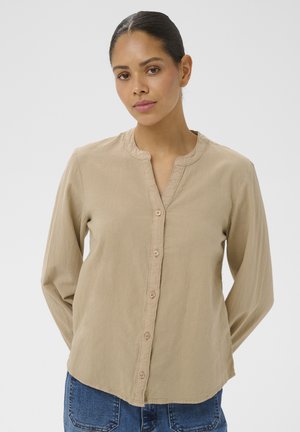 Kobieta o ciemnych włosach w beżowej koszuli zapinanej na guziki i niebieskich dżinsach, stojąca z rękami za plecami na jednolitym tle.