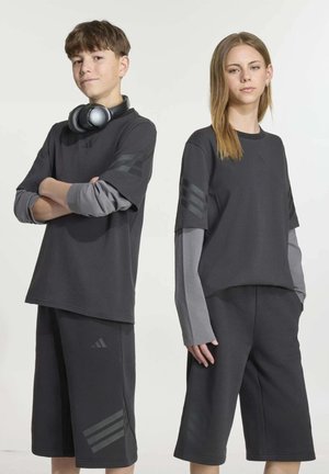 Deux adolescents présentent des tenues de sport sombres, composées de hauts superposés et de shorts aux genoux ; le garçon porte un casque audio autour du cou.