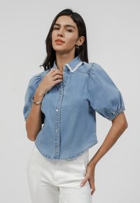 Light blue denim shirt with short puff sleeves, lace trim collar, front buttons, and a curved hemline. Worn with white pants.