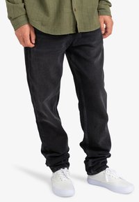 Hombre con jeans negros y zapatillas blancas, de pie con las manos relajadas a los lados, vistiendo una camisa verde con las mangas arremangadas.