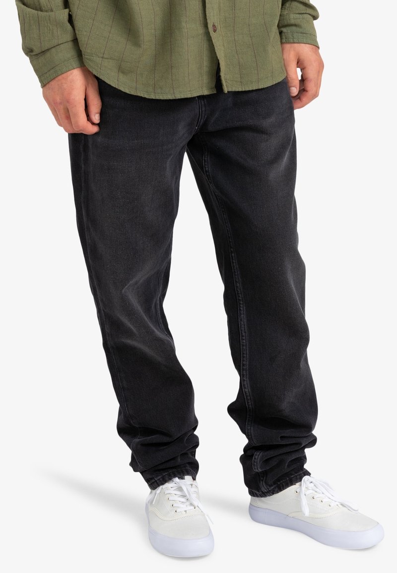 Hombre con jeans negros y zapatillas blancas, de pie con las manos relajadas a los lados, vistiendo una camisa verde con las mangas arremangadas.