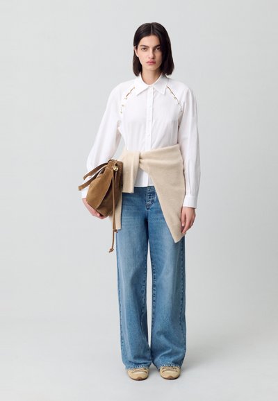 Una modella si trova di fronte a uno sfondo chiaro, indossando una camicia bianca, jeans larghi e tenendo una borsa marrone, con un maglione beige annodato in vita.