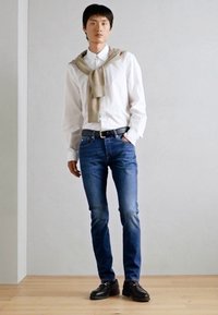 Weißes Hemd mit Knöpfen, hellbeige Pullover lässig über die Schultern gelegt, blaue Jeans, schwarze Schuhe, auf einem Holzboden stehend vor einer schlichten Wand.