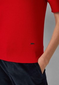 Czerwony sweter z dzianiny z krótkimi rękawami, z ribbed hem oraz małym czarnym logo na dole. W zestawie z ciemnymi, teksturowanymi spodniami.