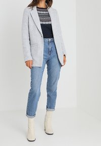 Helles graues, strukturiertes Blazer über einem navy-blauen, gemusterten Pullover, kombiniert mit hoch taillierten blauen Jeans und cremefarbenen Stiefeletten.