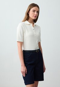 Polo bianco a coste con maniche corte, dotato di colletto e tre bottoni. Indossato con pantaloni corti in denim scuro e cintura nera.