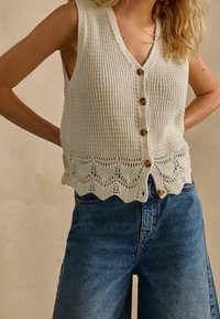 Weißer, gestrickter Weste mit V-Ausschnitt, einem strukturierten Muster, Holzknöpfen und einem gewellten Saum. Kombiniert mit blauen, hoch taillierten Jeans.